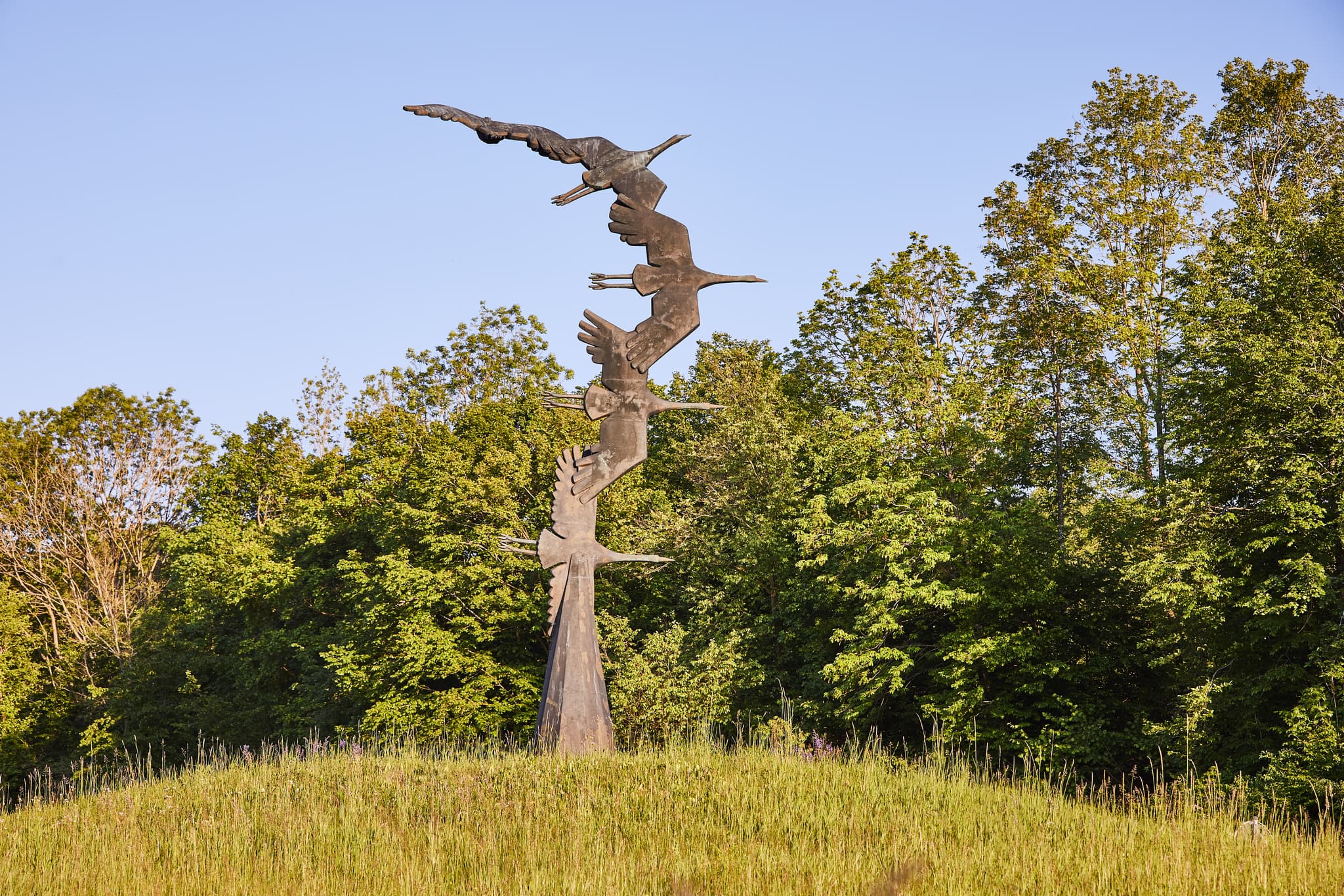 Abfahrt Kraniche Hohenwart, Altötting, Oberbayern - Kranich-Skulptur auf grasbewachsenem Hügel in Hohenwart, Emmerting, Altötting, Oberbayern. Fliegende Kraniche als Motiv in der Region Inn-Salzach, Deutschland.