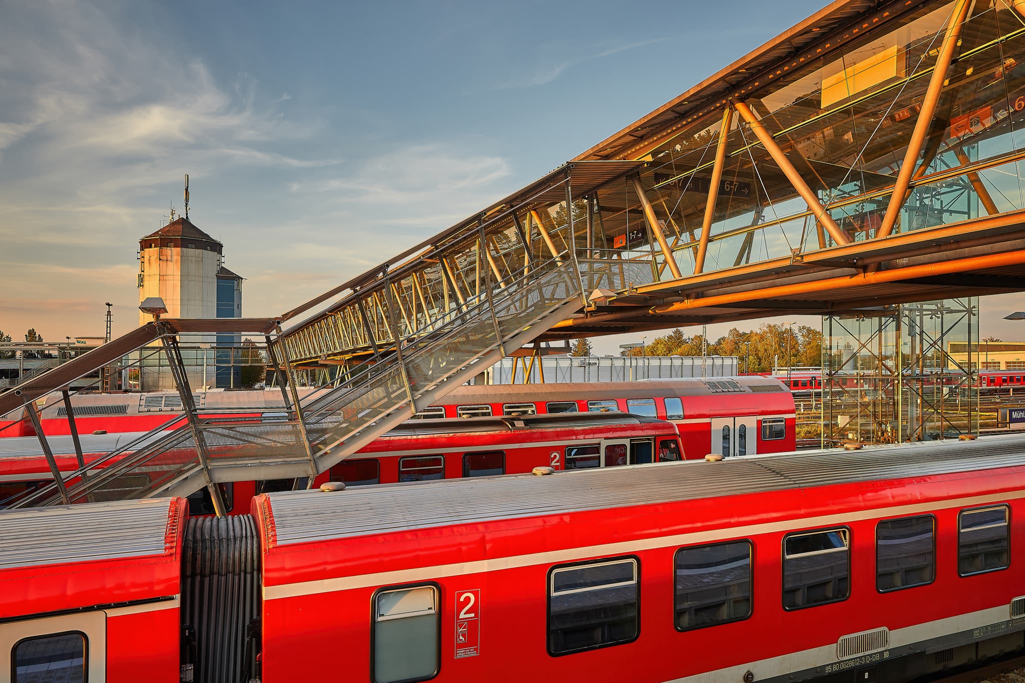Bahnhof Treppenabgang, Mühldorf am Inn, Oberbayern - Bahnhof Treppenabgang in Mühldorf am Inn, Oberbayern. Die Glasbrücke überspannt die Bahngleise mit roten Zügen. Ein Blick auf die regionale Bahninfrastruktur.