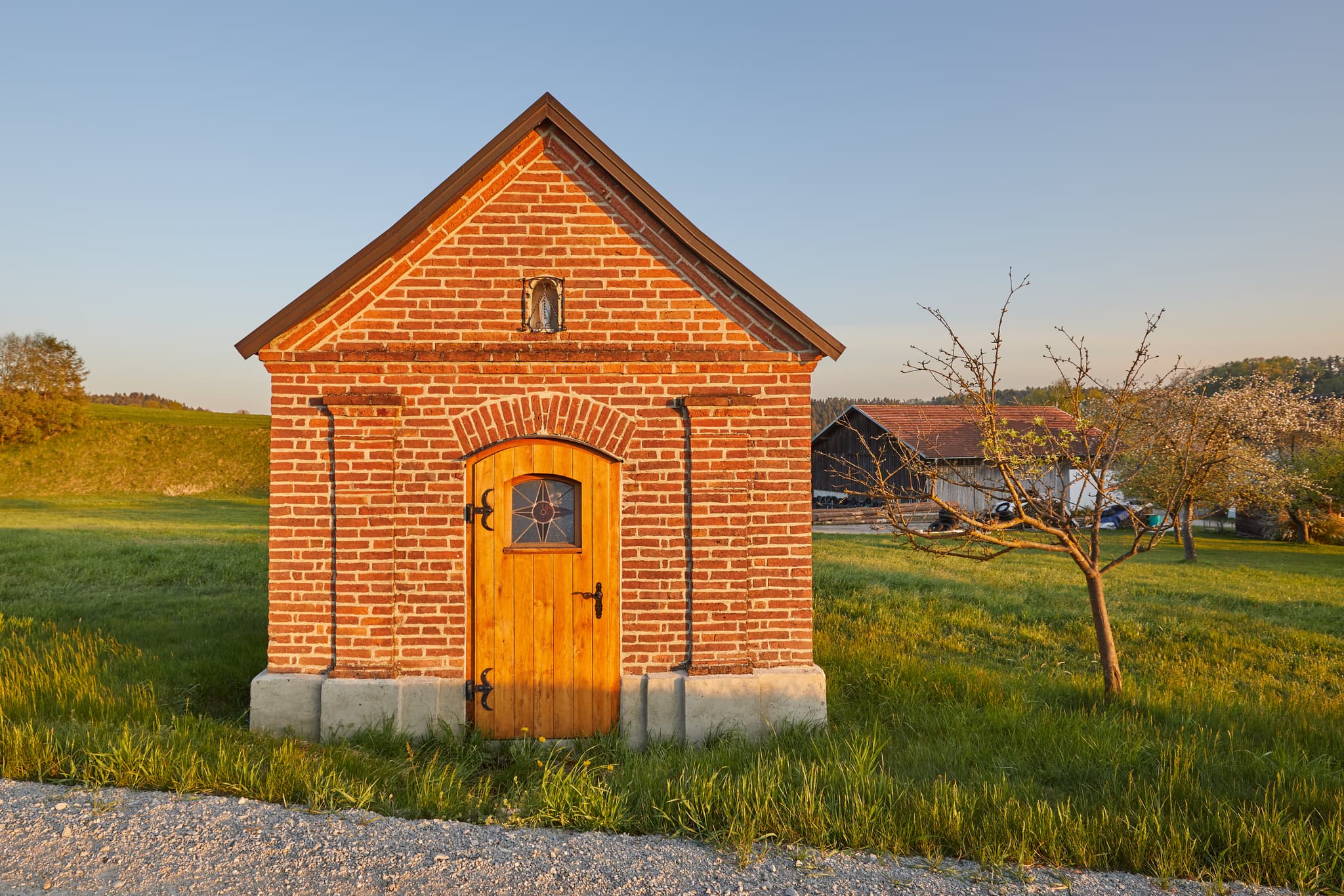 Kapelle an der Tanner Straße nach Schildthurn, Niederbayern. - Kleine Backsteinkapelle im Abendlicht in Schildthurn bei Zeilarn, Landkreis Rottal-Inn, Niederbayern, Deutschland. Umgeben von grünen Wiesen und Bäumen.