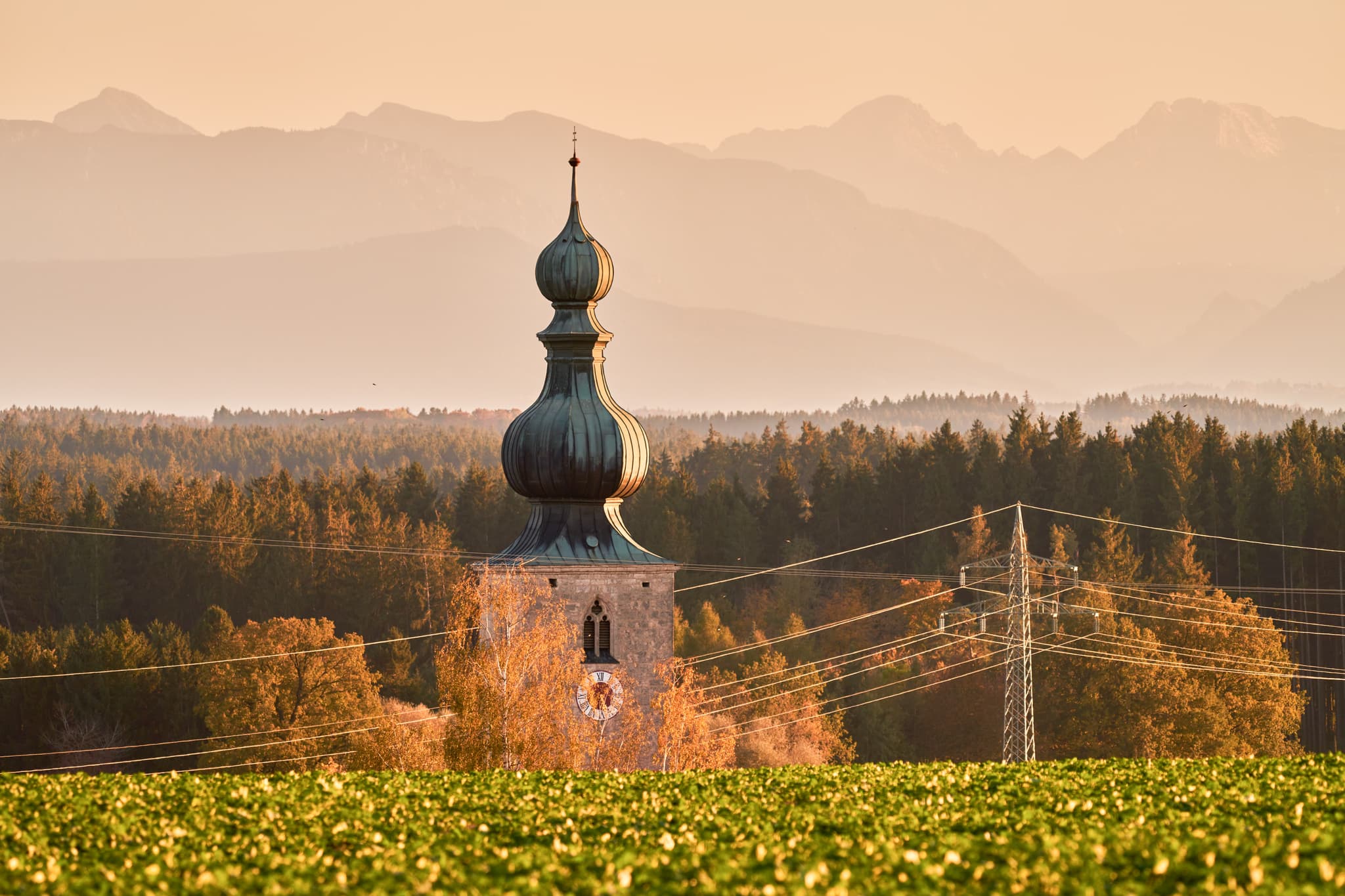 Linde Aussicht, Rainbichl, Altötting, Oberbayern - Abendliche Szene mit Zwiebelturm-Kirche in Rainbichl, Tyrlaching, Landkreis Altötting, Oberbayern, Inn-Salzach, Deutschland. Felder, Wälder, Berge, Strommast.