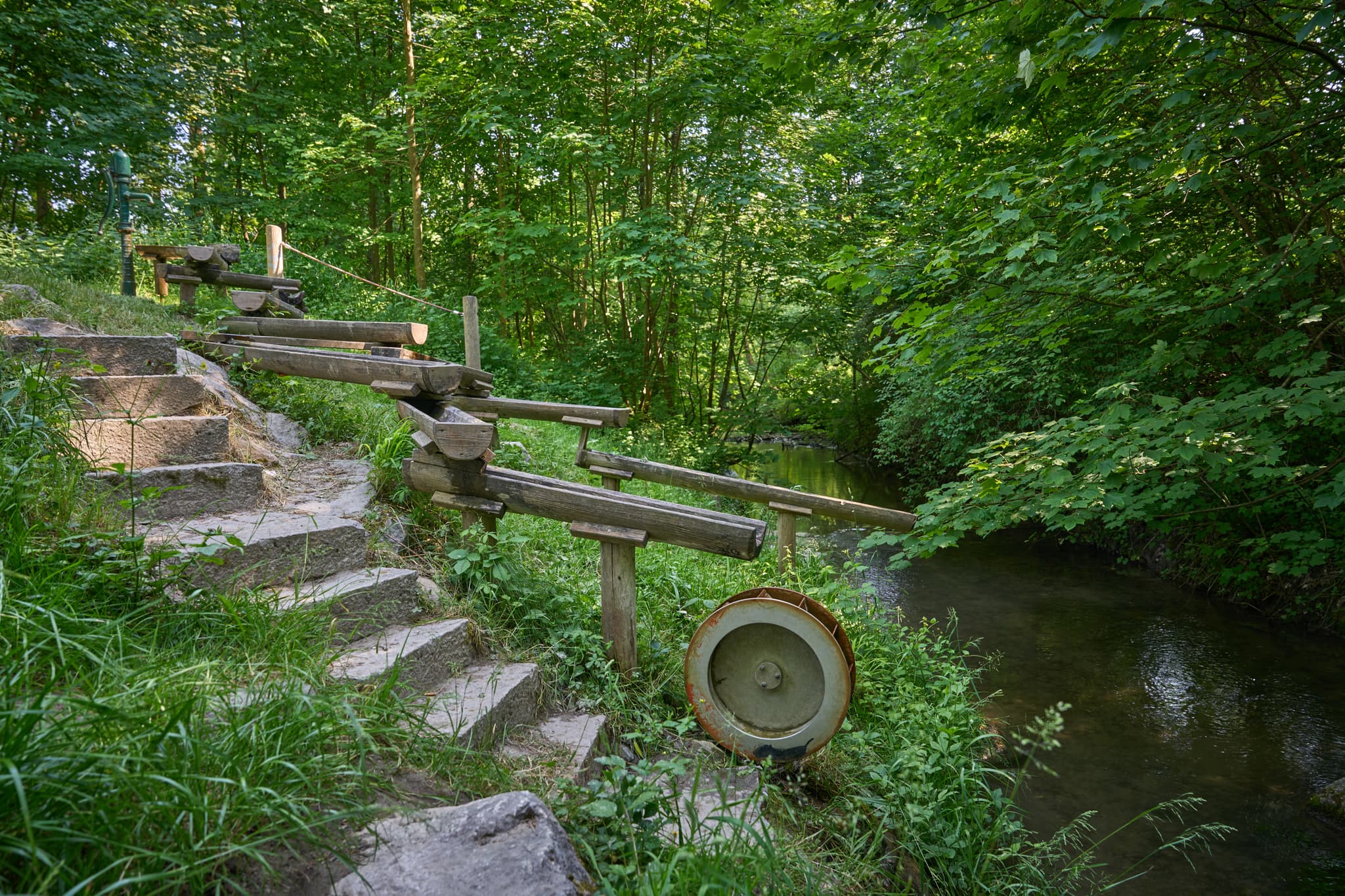 Naturerlebnisweg Innaue, Mühldorf, Speilplatz, Inn-Salzach - Idyllische Szene Naturerlebnisweg Innaue in Mühldorf am Inn, Oberbayern: Wasserspiel aus Holz am Bachlauf, Steinstufen in grüner Natur, Region Inn-Salzach.