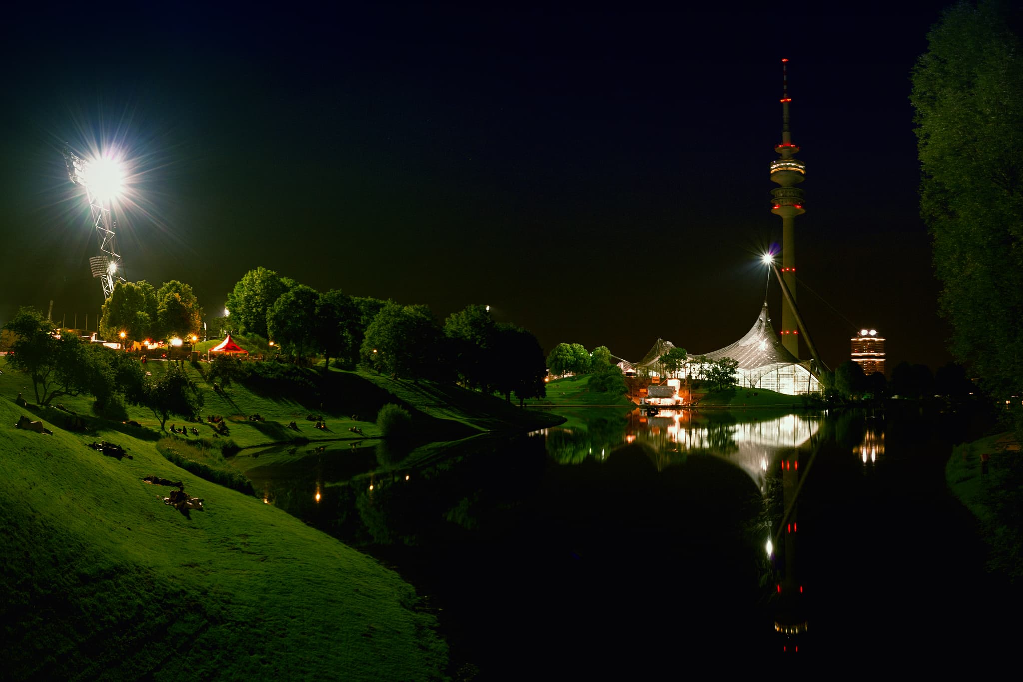 Olympiasee Nachtaufnahme Olympiaturm, München, Oberbayern - Nächtliche Ansicht des Olympiasees in München, Oberbayern, Deutschland. Beleuchtete Kulisse der Stadt mit markantem Olympiaturm. Reflexionen im Wasser.