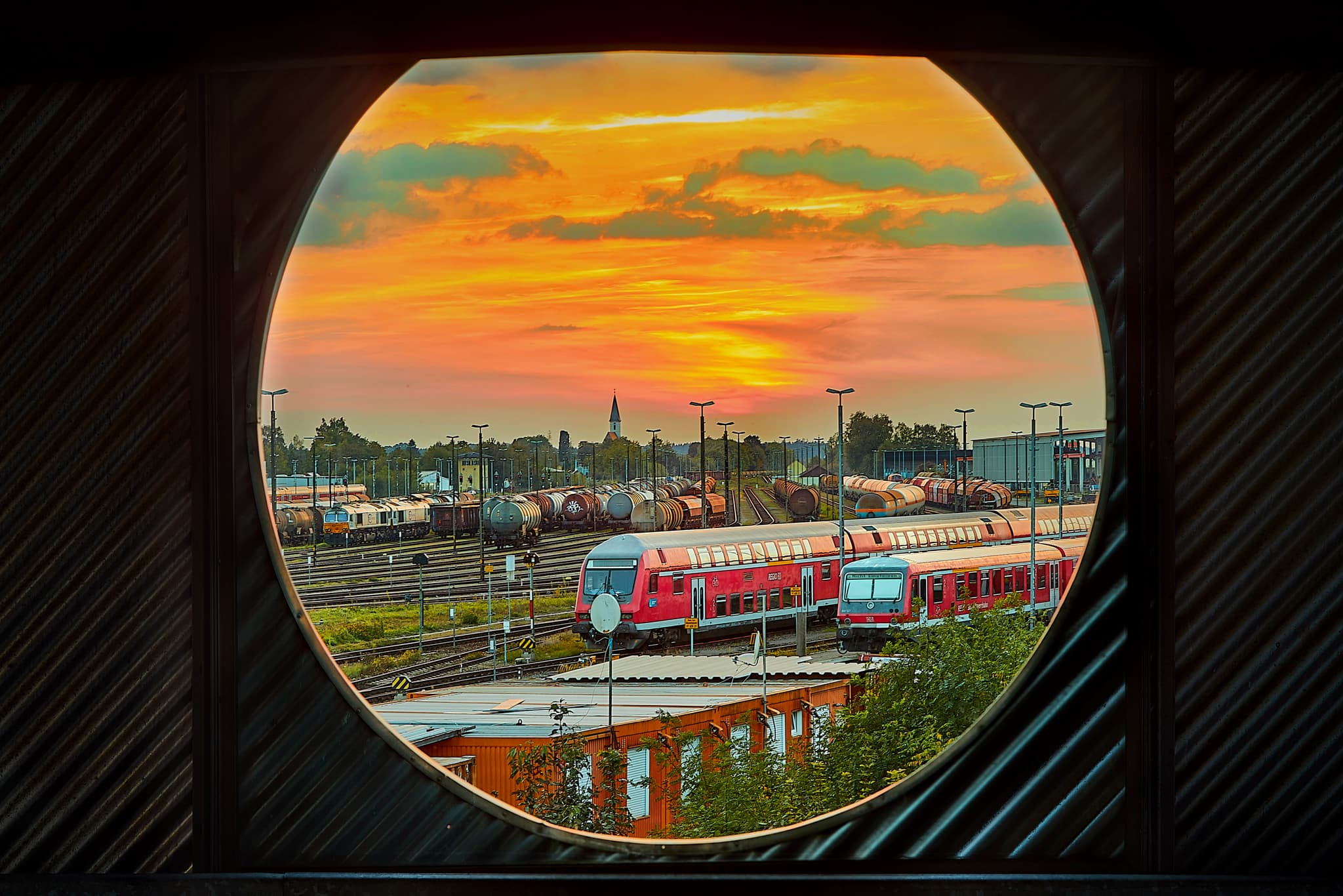 Parkhaus Fenster, Bahnhof Mühldorf am Inn, Oberbayern - Blick durch ein Fenster auf den Bahnhof Mühldorf am Inn. Zuggleise und Gebäude des Paarkhauses unter Abendrot.