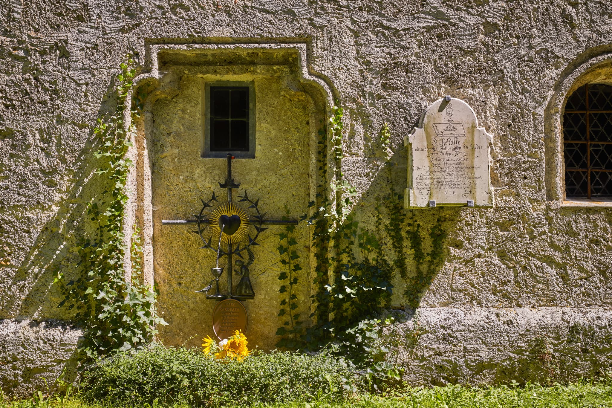 Pfarrkirche Radegundis Firedhof, St. Radegund, Braunau - Kirchenmauer mit kunstvollem Metallkreuz und Steintafel. Aufnahme aus St. Radegund, Landkreis Braunau am Inn, Innviertel, Oberösterreich, Österreich.