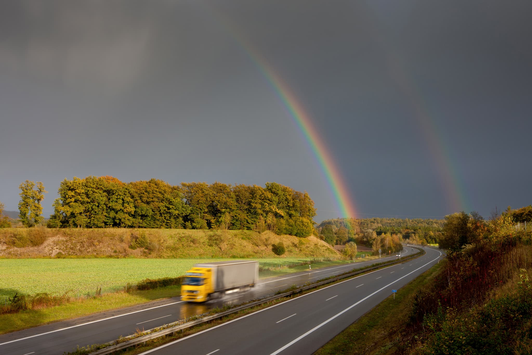 Regenbogen über der Autobahn A94 bei Mittling, Altötting - Ein Regenbogen spannt sich über die Autobahn A94 bei Mittling im Landkreis Altötting, Oberbayern. Ein LKW fährt auf der regennassen Autobahn.