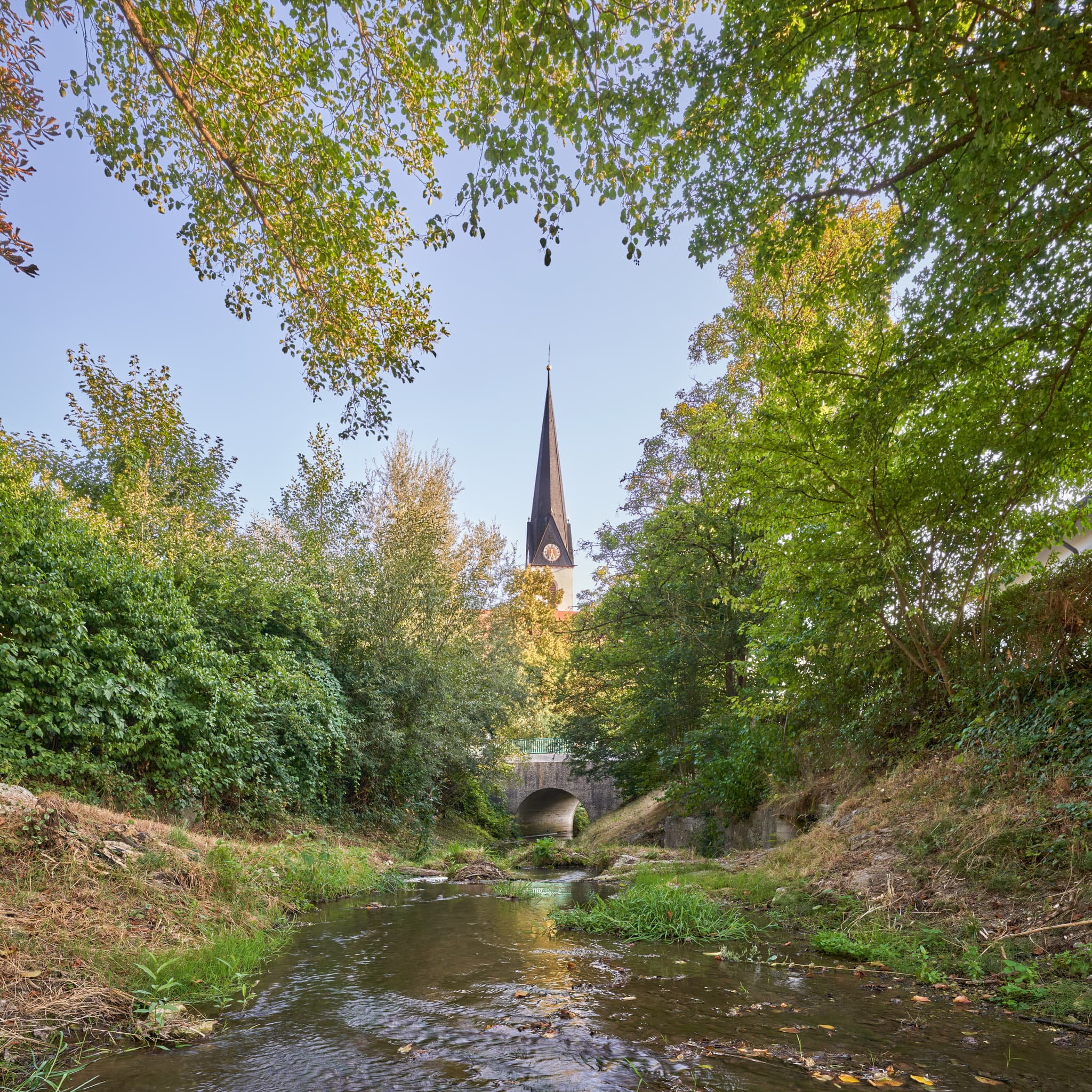 Reischach Bach am Kindergarten, Kirche, Landkreis Altötting - Der Reischachbach, Altötting, Oberbayern, vor der Brücke Erlbacher Straße. Im Hintergrund der Kirchturm St. Martin, umgeben von dichtem Grün.