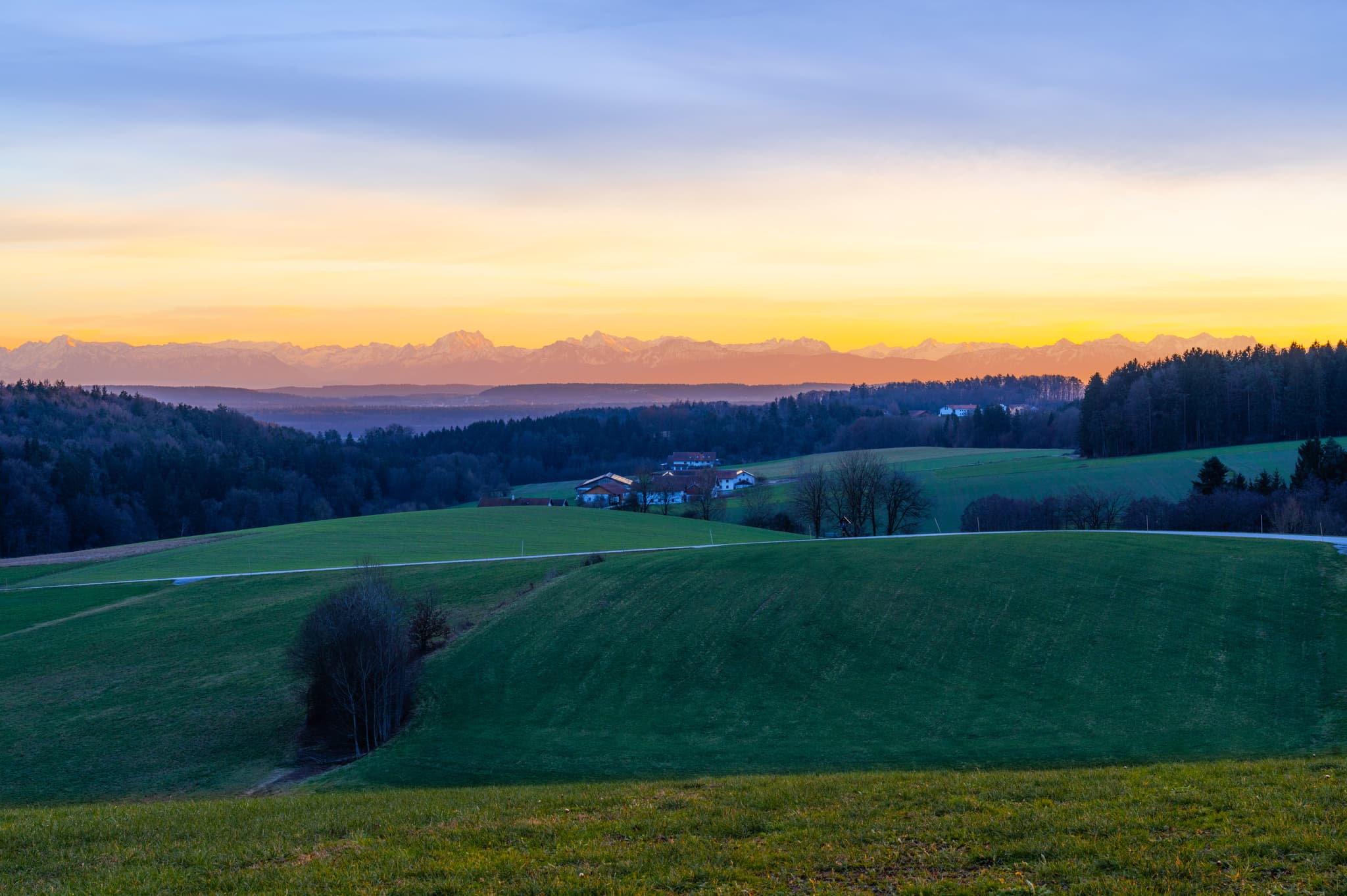 Richtung Pomming, Haizing, AÖ, Oberbayern, Inn-Salzach - Landschaft Haizing, Erlbach (AÖ), Deutschland. Grüne Hügel, Felder, Wälder Richtung Pomming. Alpen am Horizont unter Dämmerungshimmel. Ruhige ländliche Idylle.