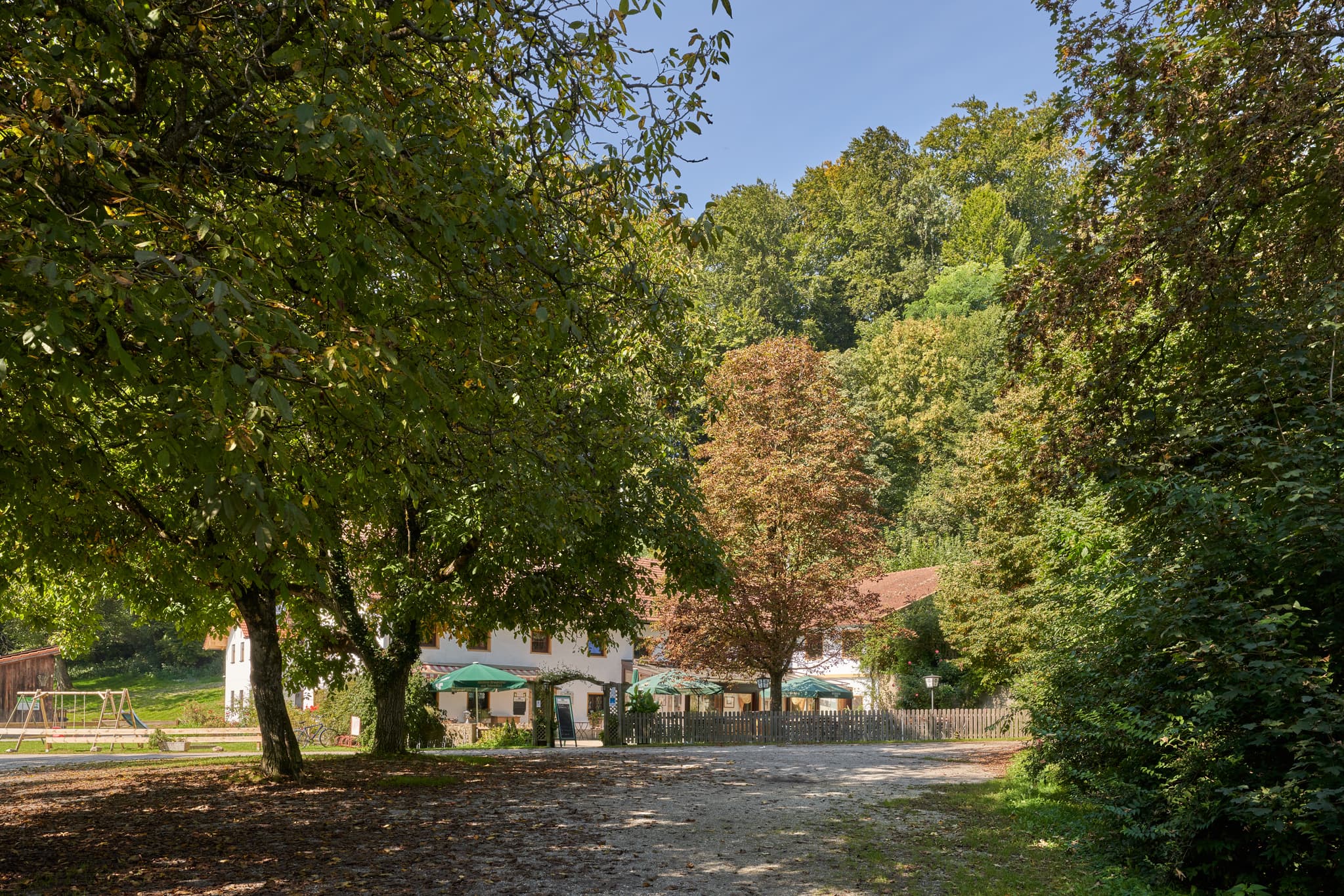 Tiefenau bei Burghausen im Landkreis Altötting, Oberbayern - Gasthaus in Tiefenau, Burghausen, Landkreis Altötting, Oberbayern. Gebäude inmitten grüner Bäume, einen sonnigen Weg und einen Spielplatzbereich.