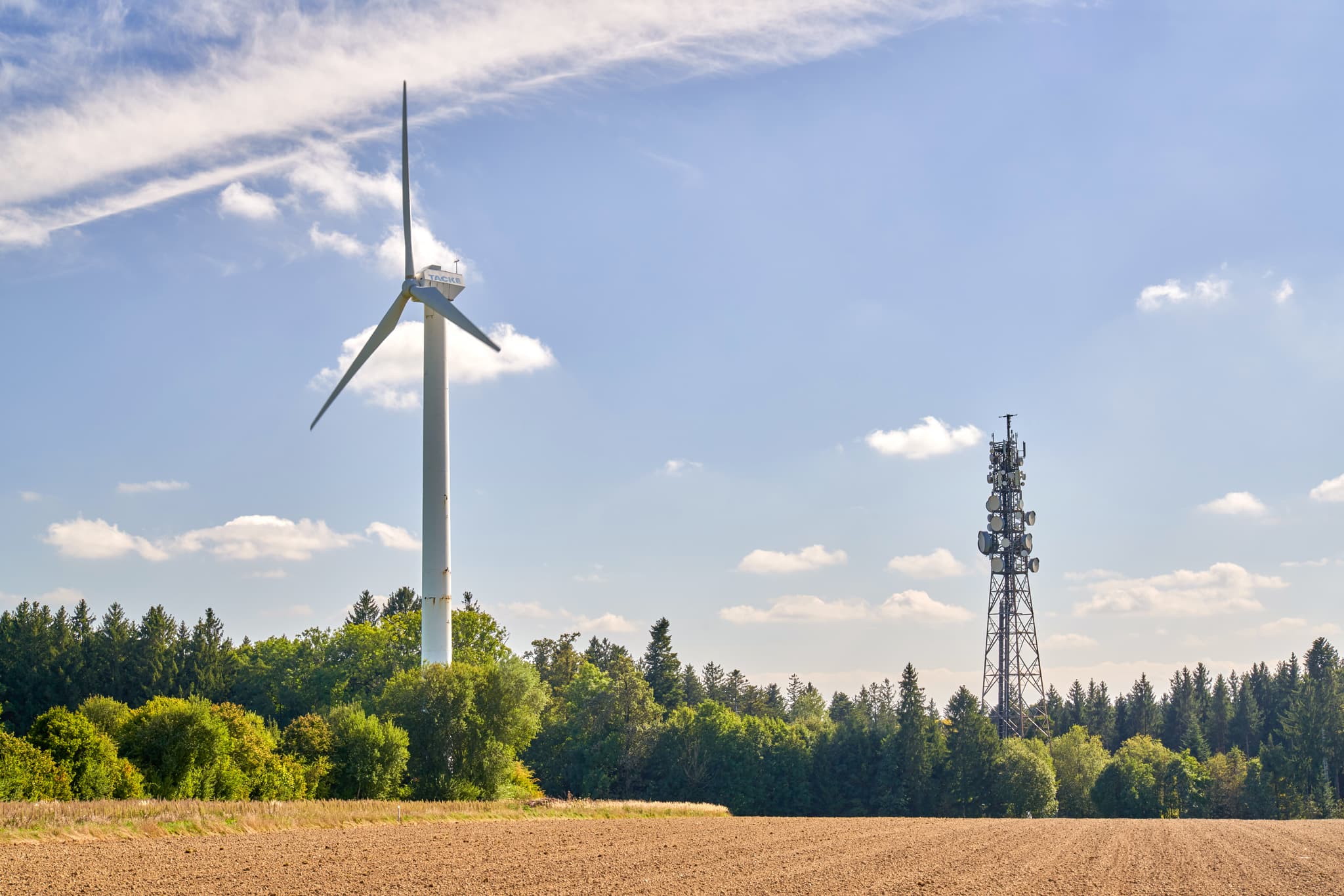 Windrad in Schnaitsee, Landkreis Traunstein, Oberbayern - Windkraftanlage und Sendemast auf Feld vor Waldhügeln unter blauem Himmel mit Wolken. Standort Schnaitsee, Traunstein, Oberbayern, Chiemgau, Deutschland.